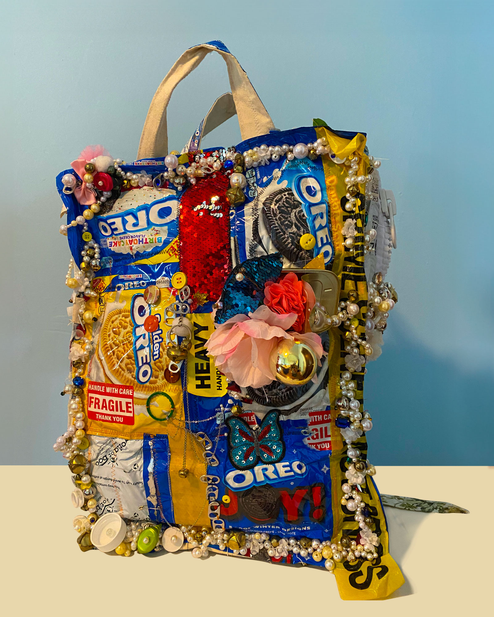 This backpack is crafted from discarded snack packages and found objects, adorned with sequins, pearls, crystals, and other materials to create a "haute couture" aesthetic. Its design aims to address issues of trash pollution and overconsumption, not by reimagining unwanted materials but by incorporating elements that draw attention to environmental concerns.