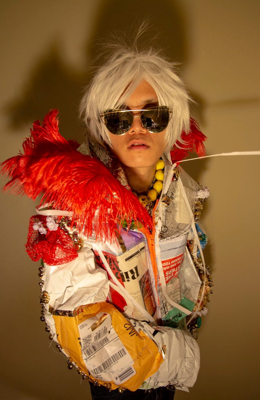 This jacket is made from newspapers, magazines, packaging, plastic, and other discarded and found materials, addressing the decline of the publishing industry due to new online business models, economic crises, and increased overconsumption, as well as trash and ocean pollution. The addition of gold jingle bells, ostrich feathers, and metallic objects enhances the jacket's haute couture aesthetic.